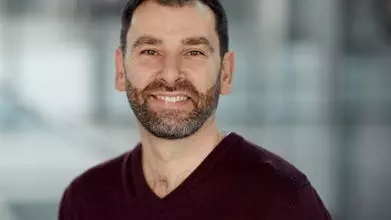 Richard Kemble has short brown hair and a short beard. He is wearing a dark v-neck jumper