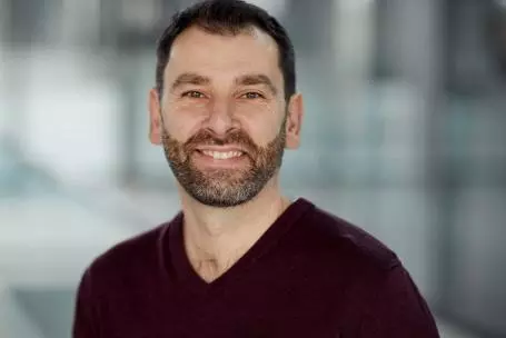 Richard Kemble has short brown hair and a short beard. He is wearing a dark v-neck jumper