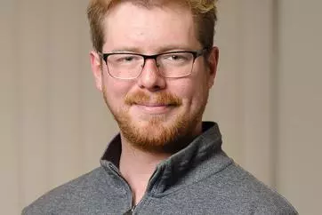 Roan Batten-Powell has short fair hair. He is wearing rectangular glasses and a grey zip-up jumper